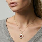 Close-up of a person wearing a gold necklace with a red stone pendant on a neutral background