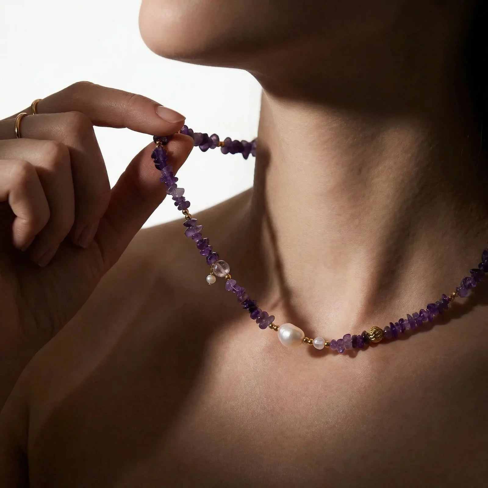 Person wearing a beaded necklace with purple amethyst beads and a pearl, against a neutral background.