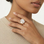 Close-up of a hand wearing a gold ring with a large baroque pearl-like stone, against a neutral background.