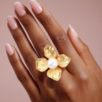 Gold flower-shaped ring with a pearl on a hand against a beige background