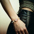 Hand wearing a bracelet with a pink stone and gold band, against a neutral background.