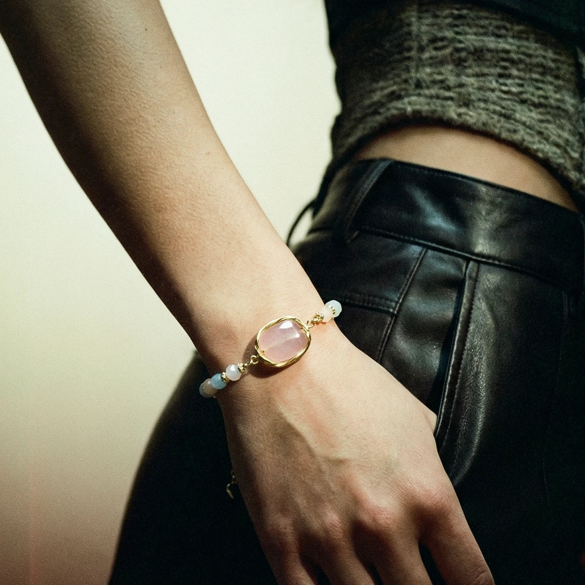 Hand wearing a bracelet with a pink stone and gold band, against a neutral background.