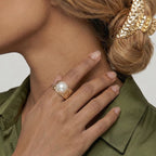 Close-up of a hand wearing a textured gold ring with a pearl, touching the neck, against a neutral background.