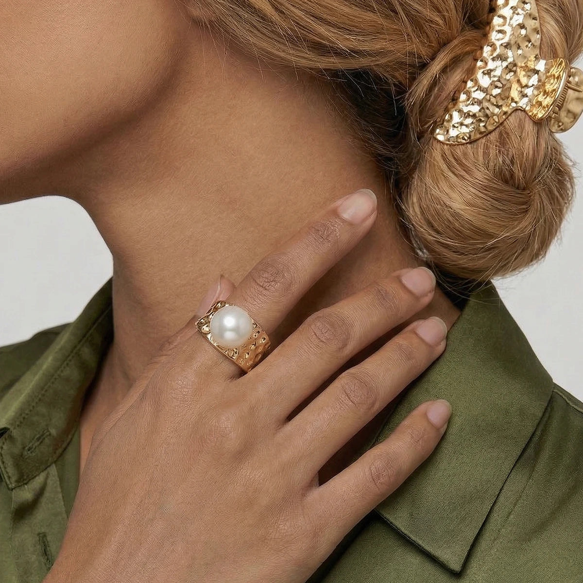 Close-up of a hand wearing a textured gold ring with a pearl, touching the neck, against a neutral background.
